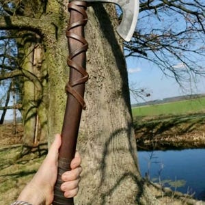 May include: A handcrafted axe with a polished steel blade and a dark wooden handle wrapped with brown leather. The axe is held in a hand, with a tree and a body of water in the background.