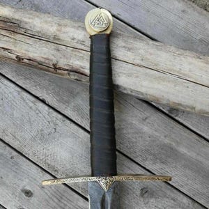 May include: A close-up of a medieval sword with a black leather-wrapped handle and a gold-colored crossguard. The pommel features a gold-colored emblem. The blade is dark and appears to be made of steel. The sword rests on a wooden surface.
