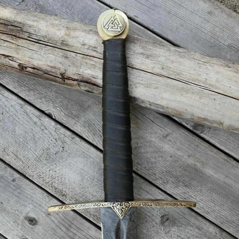May include: A close-up of a medieval sword with a black leather-wrapped handle and a gold-colored crossguard. The pommel features a gold-colored emblem. The blade is dark and appears to be made of steel. The sword rests on a wooden surface.