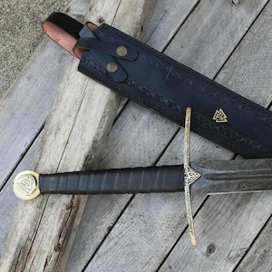 May include: A medieval sword with a leather-wrapped handle and a gold-colored pommel and crossguard. The blade is dark with a patterned design. The sword is next to a black leather sheath with decorative stitching and brass-colored buttons.
