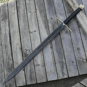 May include: A long, steel sword with a black handle and gold accents. The blade has a detailed pattern. The sword is lying on a weathered wooden surface. The pommel has a circular design.