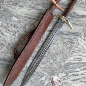 May include: A long, ornate sword with a patterned blade and a gold-colored crossguard. The handle is wrapped in dark brown leather. The sword rests in a brown leather sheath with a shoulder strap. The overall design suggests a medieval or fantasy theme.