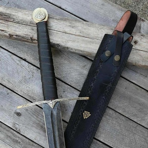 May include: A detailed image of a sword with a black handle and a gold-colored crossguard. The blade has a patterned design. A black leather sheath with gold accents is next to the sword. The sword's pommel has a gold-colored emblem.