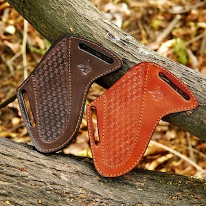 Puede incluir: Dos fundas de cuero para cuchillos en tonos marrón y naranja. Las fundas tienen un patrón de tejido de cesta y un pequeño emblema. Están sobre una rama de árbol.