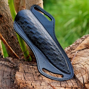 May include: A black leather knife sheath with a decorative embossed pattern. The sheath has a loop for a belt and is designed to hold a knife. The sheath is shown on a tree stump.