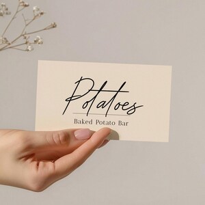 May include: A rectangular cream-colored card featuring the black text "Potatoes" in a cursive script font, with the words "Baked Potato Bar" printed in a smaller, simple sans-serif font directly underneath the main title.