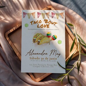 Op de afbeelding: Een rechthoekige uitnodigingskaart met de tekst "TACO 'BOUT LOVE" en illustraties van taco's en maracas. De kaart bevat de naam "Alexandra May" en evenementdetails. De kaart ligt op een houten dienblad, met een roze en blauwe achtergrond.