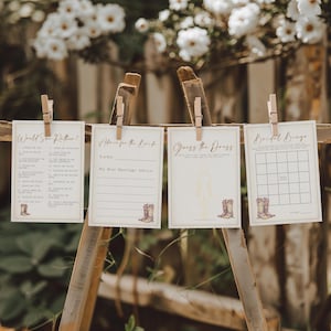 Puede incluir: Cuatro tarjetas de juegos de boda, incluyendo "Would She Rather?", "Advice for the Bride", "Guess the Dress" y "Bridal Bingo". Cada tarjeta está sujeta por una pinza de madera en una estructura de madera, con un fondo floral.
