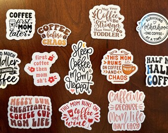 Puede incluir: Una colección de pegatinas blancas con texto negro y de colores sobre una superficie marrón. Las pegatinas tienen frases como "Coffee first, Mom later", "Coffee before chaos" y "Too much toddler, not enough coffee".