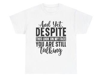 以下が含まれることがあります： 「And Yet, DESPITE THIS LOOK ON MY FACE YOU ARE STILL talking」という黒い文字がプリントされた白いTシャツ。柔らかい素材でできており、普段使いに適しています。
