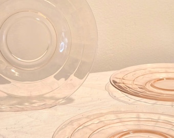 Puede incluir: Una colección de platos de vidrio transparente y rosa. Los platos tienen un diseño circular con anillos concéntricos. Los platos están dispuestos sobre una superficie blanca, con una iluminación suave que crea sombras y reflejos en el vidrio.