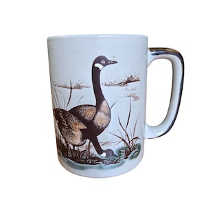 May include: A ceramic mug with a detailed illustration of two geese in a marsh setting. The geese are depicted with brown and black feathers, and the mug has a white background with a brown handle.