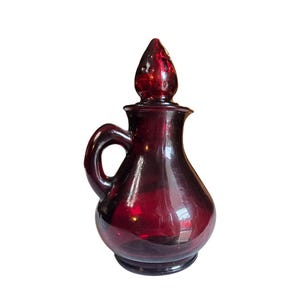 May include: A ruby red glass cruet with a handle and a pointed stopper. The cruet has a bulbous body and a small, round base. The handle is a loop shape. The stopper is a teardrop shape.