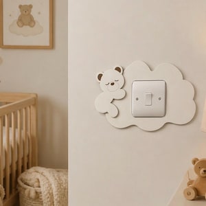 Könnte beinhalten: Eine hellbeige, wolkenförmige Lichtschalterabdeckung mit einem schlafenden Bären-Design. Die Schalterabdeckung ist an einer hellbeigen Wand in einem Kinderzimmer angebracht. Ein gerahmtes Bild eines Teddybären auf einer Wolke ist sichtbar.
