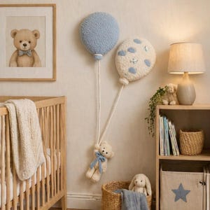 Op de afbeelding: Twee pluche wandballonnen in lichtblauw en crème met wolkenpatronen, verbonden door witte touwen aan een kleine pluche teddybeer met een blauw lint. De scène bevat een houten wieg, een ingelijste berenprint en een boekenplank met een lamp.