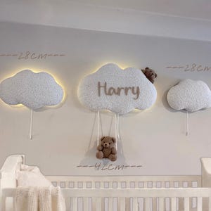 Op de afbeelding: Drie witte, wolkvormige wanddecoraties met warme verlichting. De middelste wolk heeft de naam "Harry" in bruine letters en een kleine teddybeer die eronder hangt. De wolken zijn ongeveer 28 cm breed.