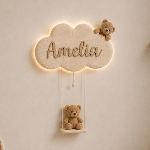 May include: A cloud-shaped wall light featuring the name Amelia in script lettering, illuminated by a warm back glow. A small teddy bear sits on a wooden swing suspended from the cloud, with another bear attached to the top right edge.