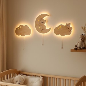 Puede incluir: Una escena de guardería con una cuna de madera y decoraciones de pared. Las decoraciones de pared incluyen tres recortes de madera iluminados: una nube, una luna creciente con un oso y otra nube, todos con acentos de estrellas. Un osito de peluche y un jarrón están en un estante.