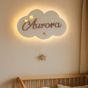 Könnte beinhalten: Ein Kinderzimmer mit einem Holzbett, einem Halbmondkissen und einem quadratischen Kissen. Darüber eine wolkenförmige Wandleuchte mit dem Namen "Aurora" und Sternen. Ein geflochtener Korb und eine Decke vervollständigen die gemütliche Einrichtung.