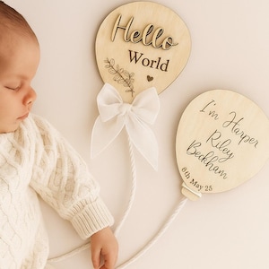 Op de afbeelding: Een pasgeboren baby in een crèmekleurige kabelgebreide outfit ligt naast twee houten ballonschilden. Op het ene bord staat "Hello World" en op het andere "I'm Harper Riley Beckham 6th May 25". Een witte strik is aan de eerste ballon bevestigd.