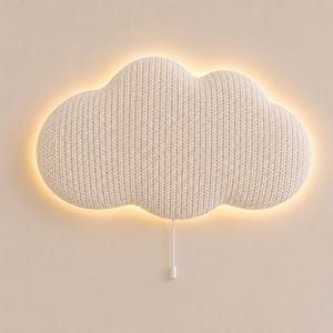 Pode incluir: Uma luminária de parede em forma de nuvem com uma capa de tecido de malha texturizada em tom off-white. A lâmpada emite um brilho quente e suave por trás das bordas, criando um efeito de halo. Um interruptor de cordão branco pende verticalmente.