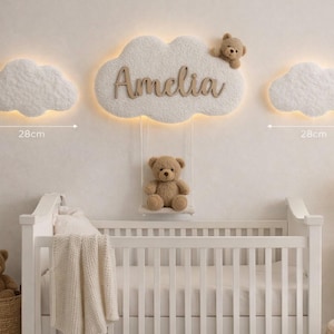May include: A nursery wall decor set featuring three fluffy cloud-shaped lights, each 11 inches wide, with the name "Amelia" in wooden letters. A teddy bear sits on a swing below the central cloud, and another in a basket. A white crib is in the foreground.