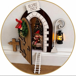May include: A decorative wooden elf door with a Christmas wreath and a sign reading "Elves Only." Inside, an elf reads a book near a Christmas tree. A small ladder and a lantern are also present.