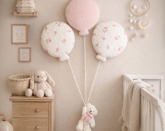 May include: Three plush balloon wall decorations in soft pink and white with floral patterns, attached to white strings held by a small white teddy bear. The set includes a solid pink balloon and two white balloons with pink flower prints.