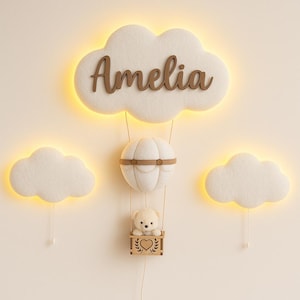 May include: A nursery wall decor set featuring a cloud-shaped light with the name "Amelia" in wooden letters. A hot air balloon with a teddy bear in a basket hangs below. Two additional cloud lights complete the set.