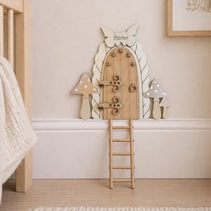 Puede incluir: Puerta de hadas de madera con una parte superior en forma de mariposa que dice "Fairies". La puerta tiene una escalera de madera, dos decoraciones de hongos y bisagras decorativas. La puerta es de madera clara.