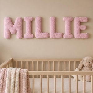 May include: A set of six large, plush, light pink letters spelling the name MILLIE, mounted on a wall above a wooden crib. The letters have a soft, textured, sherpa-like fabric finish and a rounded, three-dimensional block font style.