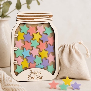 Könnte beinhalten: Holz-Stern-Glas im Einmachglas-Design, gefüllt mit bunten sternförmigen Spielsteinen. Das Glas hat einen Holzdeckel und den Text "Josie's Star Jar". Ein kleiner Beutel mit Kordelzug und lose Sterne in verschiedenen Farben liegen neben dem Glas.