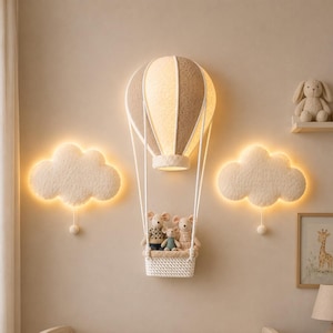 Puede incluir: Una lámpara de pared con forma de globo aerostático con dosel texturizado en beige y blanco, que incluye una cesta tejida blanca con tres pequeñas figuras de ratón de peluche. Dos lámparas de pared blancas con forma de nube iluminan los lados.