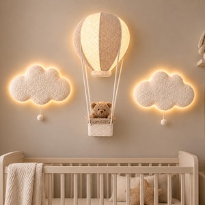 May include: A wall-mounted hot air balloon lamp with a beige and white textured canopy, featuring a small basket containing a brown teddy bear, flanked by two illuminated white cloud-shaped wall lights with hanging pull cords.