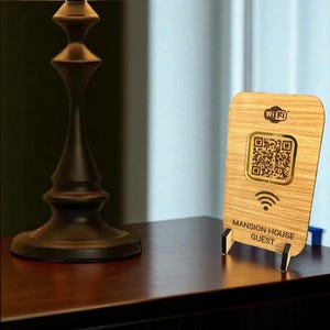 May include: A wooden WiFi sign with a QR code, WiFi symbol, and the text "MANSION HOUSE GUEST." The sign is propped up on a dark wooden surface next to a lamp with a beige lampshade. The sign is made of light-coloured wood.
