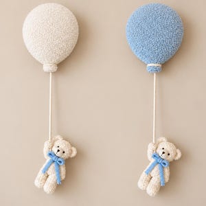 Könnte beinhalten: Zwei Wanddekorationen mit cremefarbenen Teddybären, die jeweils eine Schnur halten, die an einem strukturierten Ballon befestigt ist. Ein Ballon ist cremefarben und der andere hellblau. Jeder Bär trägt eine kleine blaue Schleife am Hals.