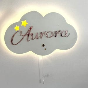 Pode incluir: Uma luz de parede em forma de nuvem branca com o nome "Aurora" em fonte cursiva. A luz tem duas estrelas amarelas e uma estrela pequena. Um cabo com um interruptor pendura-se por baixo.