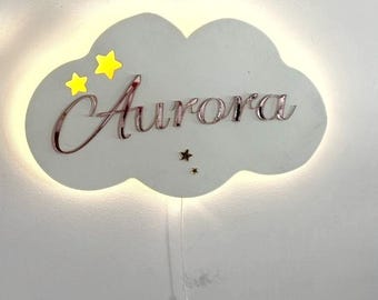 Może przedstawiać: Biała lampa ścienna w kształcie chmury z napisem "Aurora" w kursywie. Lampa ma dwie żółte gwiazdy i małą gwiazdkę. Poniżej wisi przewód z przełącznikiem.