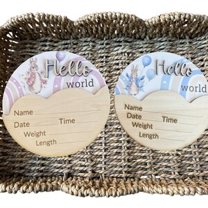 May include: Two wooden baby announcement plaques with a bunny and rainbow design. Each plaque has the words "Hello world" and space to write the baby's name, date, time, weight, and length. The plaques are in a woven basket.