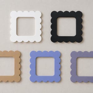 May include: Five square picture frames with scalloped edges in white, black, tan, light blue, and lavender. The frames have a square opening in the center. The frames are arranged on a neutral background.