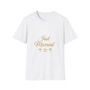 Puede incluir: Camiseta blanca con la frase "Just Married" en letras doradas. El diseño incluye dos palmeras y anillos de boda. La camiseta es de cuello redondo.