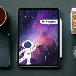 Könnte beinhalten: Ein digitales Tablet zeigt ein Notizbuchcover mit Weltraumthema. Das Cover zeigt eine Astronautenillustration vor einem lila-blauen Galaxiehintergrund. Der Text "My Notebook" befindet sich in einem weißen Oval. Ein weißer Stift liegt auf dem Tablet.