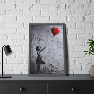May include: Framed artwork featuring a black and white silhouette of a young girl reaching for a red heart-shaped balloon. The artwork is set against a textured, distressed wall background, with a dark gray frame.