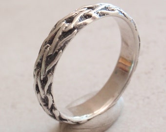 May include: A silver ring featuring a textured, braided pattern along the outer band. The metal has a polished finish on the interior, while the exterior surface displays a detailed, interwoven design with dark, recessed areas between the links.