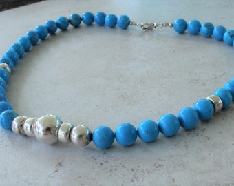 May include: A beaded necklace featuring round, bright blue turquoise-colored beads interspersed with polished silver-toned metal beads. The necklace includes a central larger silver-toned bead and a silver-toned metal lobster claw clasp closure.