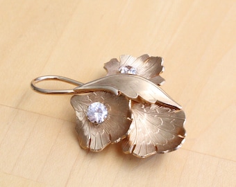 May include: A gold-toned metal brooch shaped like three ginkgo leaves with textured, fan-like surfaces. Two of the leaves are centered with a single, round, clear faceted gemstone. The brooch features a curved stem extending from the top.