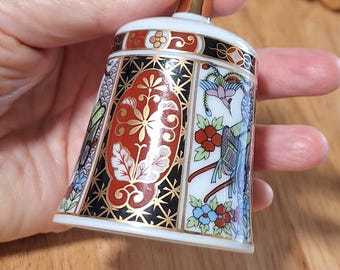 May include: A porcelain bell featuring a gold-colored handle and a white base decorated with intricate panels of red, black, and blue floral and bird motifs. The design includes geometric patterns and gold accents throughout the surface.