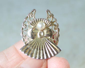 May include: A silver-toned metal brooch shaped like an angel with filigree wings, a smooth circular head, and a pleated fan-like skirt. A single small white pearl is set at the center of the angel's chest, just below the head.