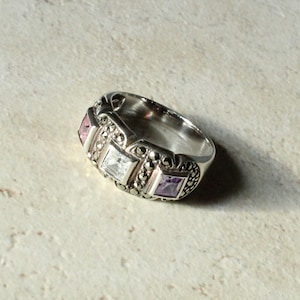May include: A silver ring with three square gemstones in pink, white, and purple. The ring has a decorative band with small, round accents. The ring is set against a neutral background.