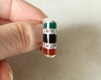 May include: A silver ring featuring three horizontal inlaid stone bands in green, black, and red, separated by two thin rows of small, sparkling marcasite stones. The ring has a polished metallic finish and a curved, rectangular face design.